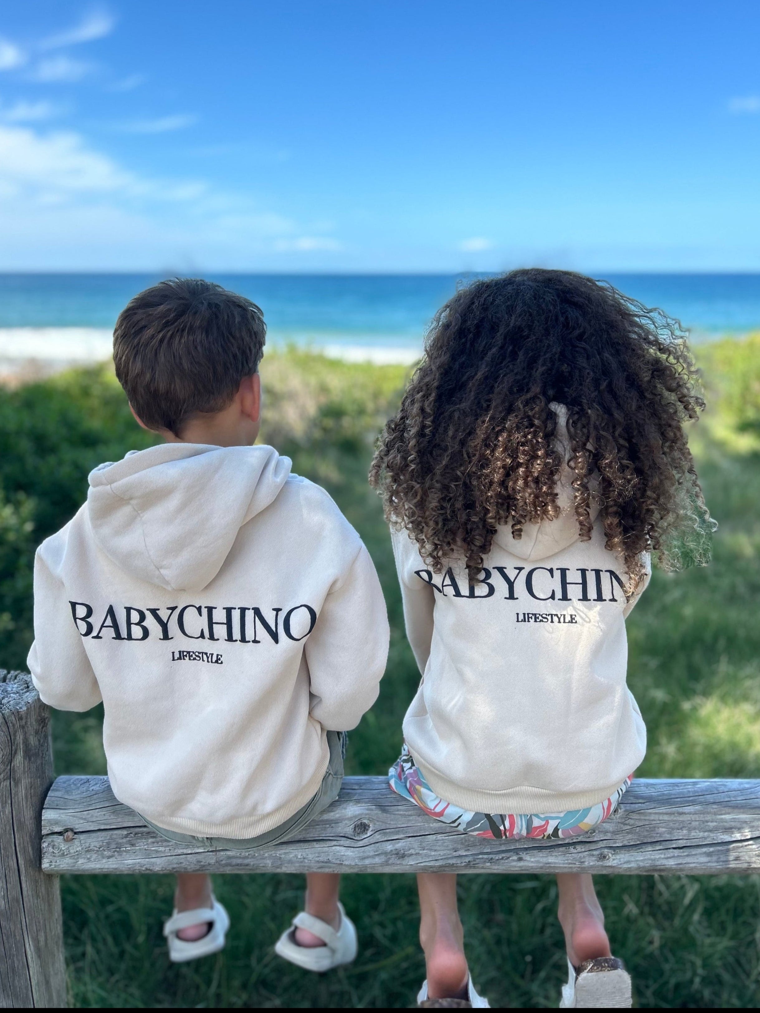 Two children wearing BABYCHINO hoodies sitting on a wooden bench by the ocean.